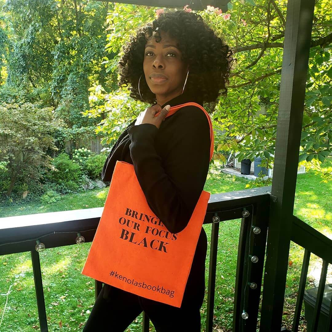 A Black woman dressed in black carrying Kenola's Bookbag's signature orange cotton tote bag with the phrase "Bringing Our Focus Black".