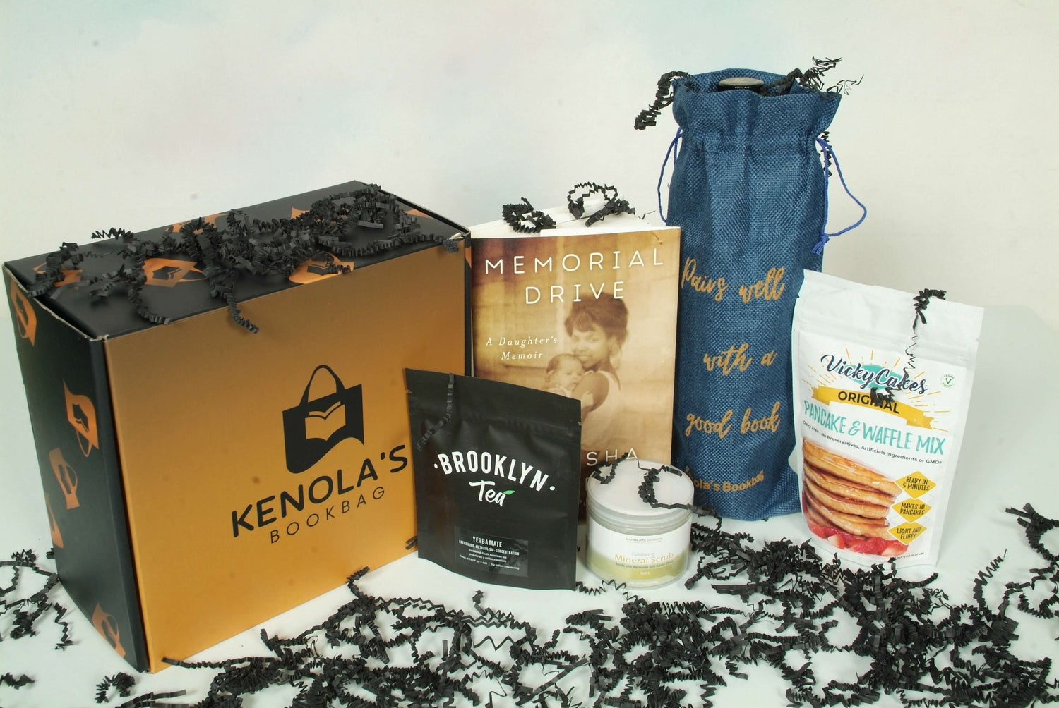 A Kenola's curated subscription box surrounded by a book written by a Black author, a wine bag, a mineral body scrub, pancake and waffle mix, and a bag of loose tea. All from Black owned businesses. Decorated with black crinkle paper.