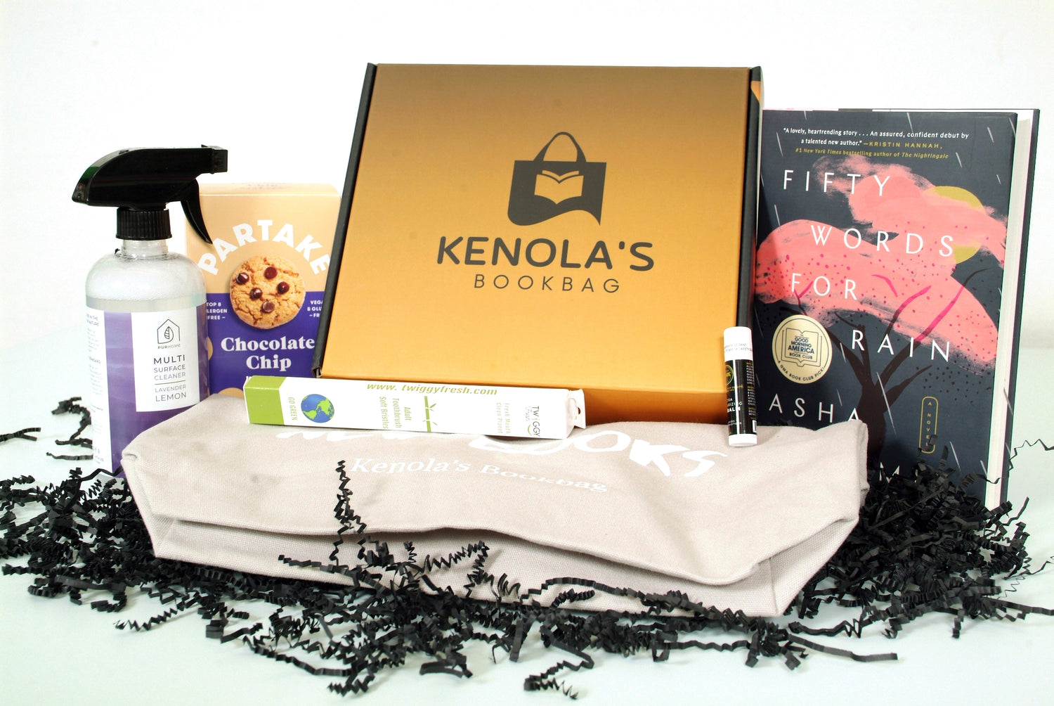 A Kenola's curated subscription box surrounded by a book written by a Black author, a cotton crossbody bag, a home cleaning spray, chocolate chip cookies, and a toothbrush.  All from Black owned businesses. Decorated with black crinkle paper.   